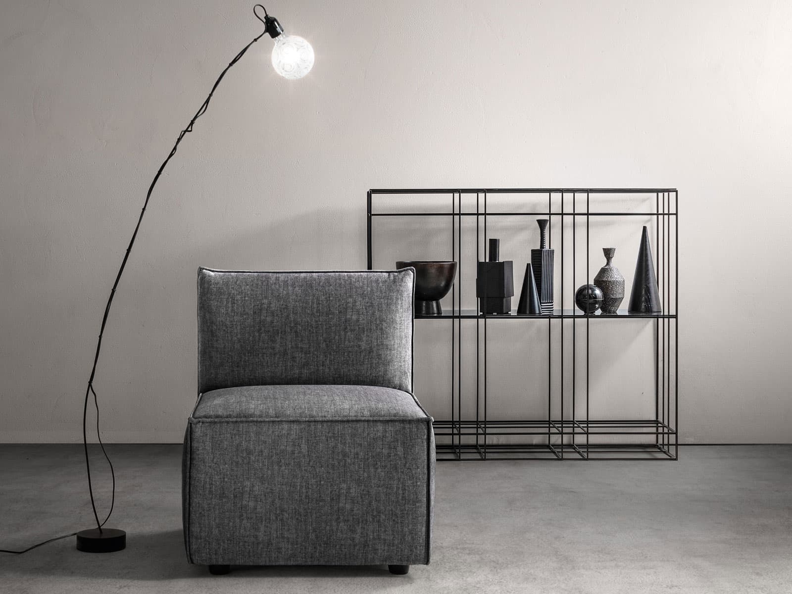 Lampada da terra ad arco sottile con sfera in vetro accanto a una poltroncina grigia e libreria in metallo