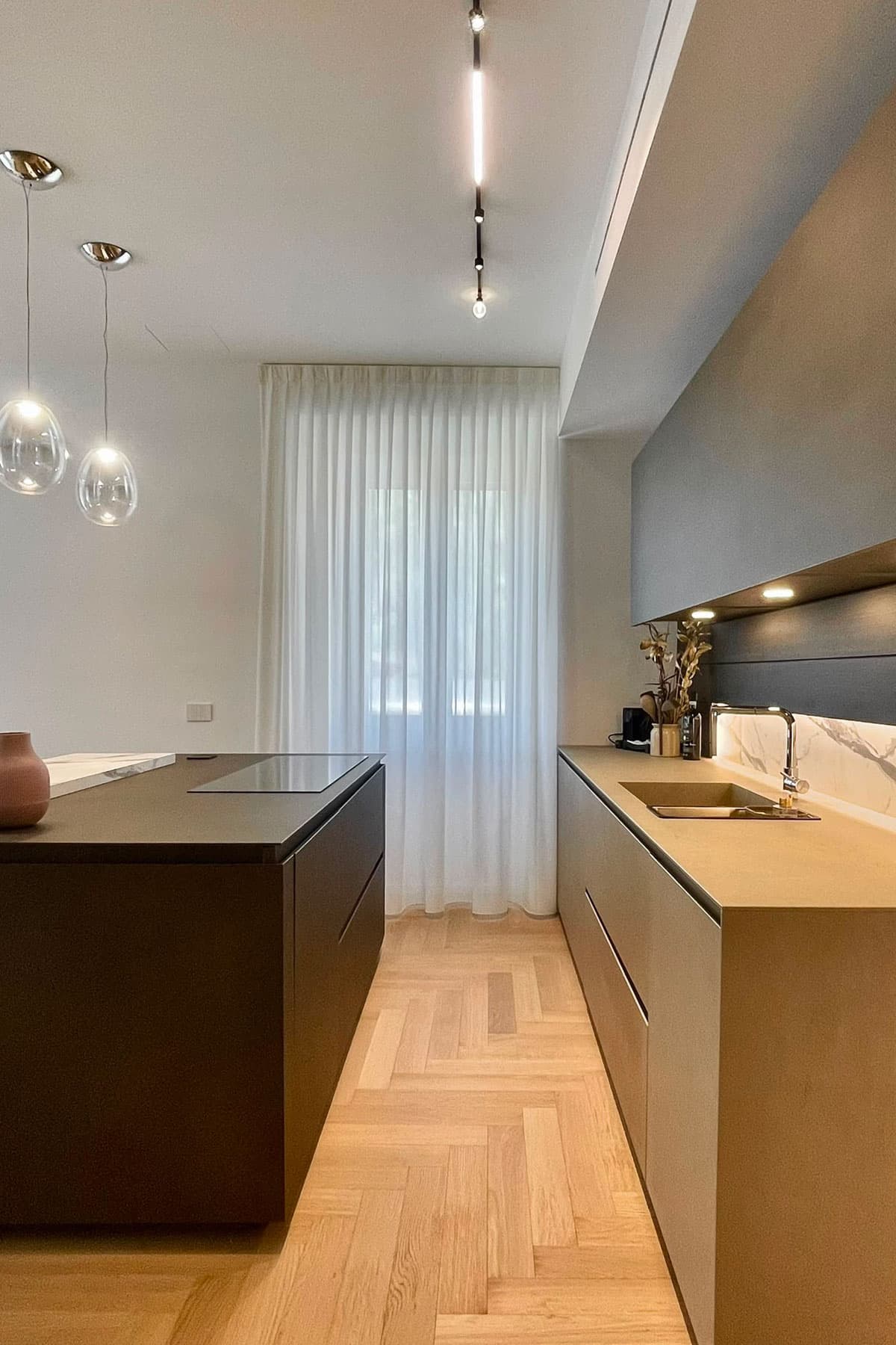 Isola scura con piano cottura e cucina lineare in gres, binario luci e parquet a spina
