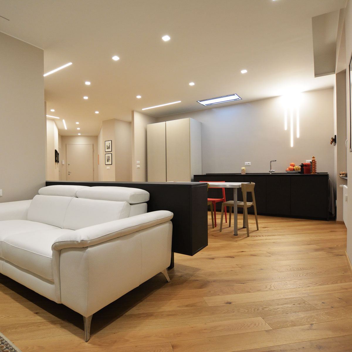 Open space con divano in pelle bianca, madia scura divisoria, cucina nera e tagli di luce a soffitto